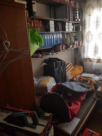 Überfülltes Arbeitszimmer mit Regalen voller Aktenordner, Werkzeug und Taschen im Chaos.