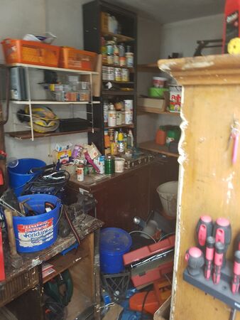 Unordentliche Werkstatt mit Werkzeugen, Regalen, Eimern und diversen Arbeitsmaterialien.